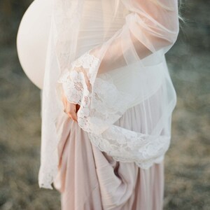 May include: A pregnant person wearing a white lace dress with a sheer overlay. The dress has a flowing skirt and a fitted bodice. The person is standing in a field of tall grass.