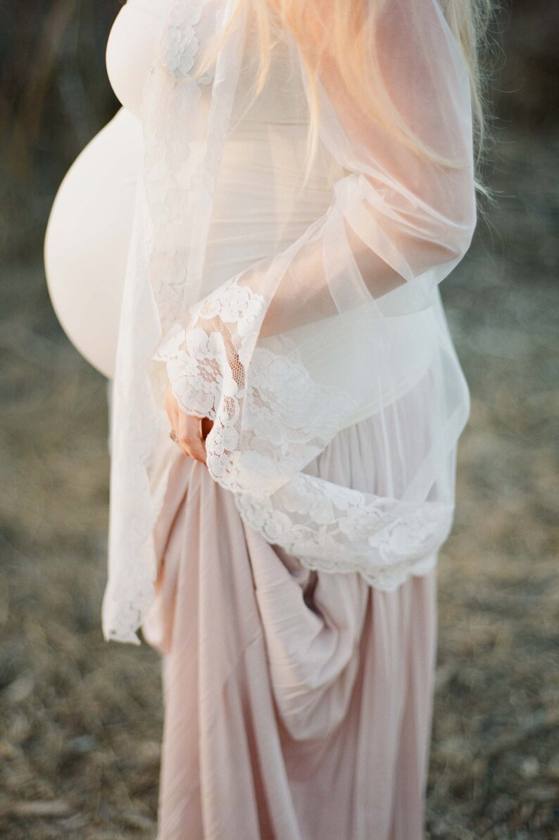 May include: A pregnant person wearing a white lace dress with a sheer overlay. The dress has a flowing skirt and a fitted bodice. The person is standing in a field of tall grass.