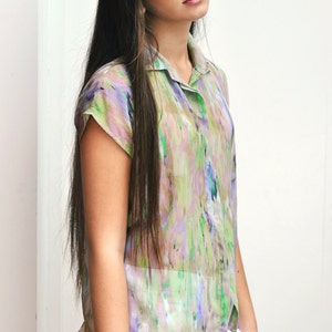 Könnte beinhalten: Kurzärmliges Hemd mit einem Aquarellmuster in Grün-, Lila- und Rosatönen. Das Hemd wird mit weißen Shorts kombiniert. Das Model hat lange, dunkle Haare.