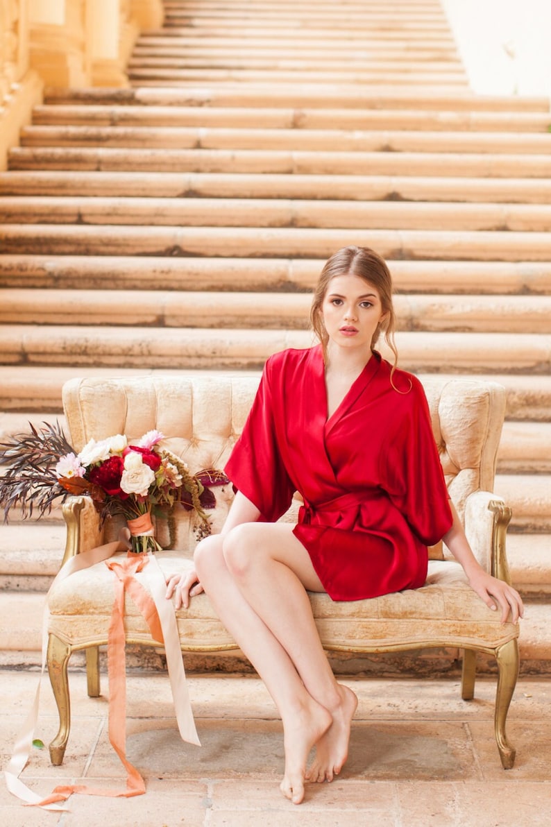May include: A woman wearing a red silk robe sits on a vintage white couch with a bouquet of flowers in her lap. The couch is in front of a set of stairs.