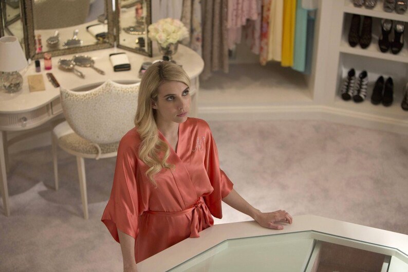 May include: A woman in a coral satin robe stands in a luxurious walk-in closet. The closet is filled with clothes and shoes, and the woman is looking up at something.