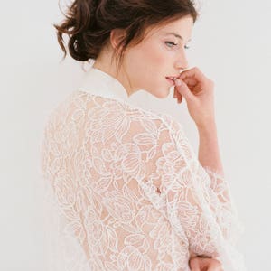 Op de afbeelding: Een witte kanten robe met een bloemenpatroon. De robe is doorschijnend en heeft lange mouwen. Het model draagt de robe en heeft haar haar in een knotje.