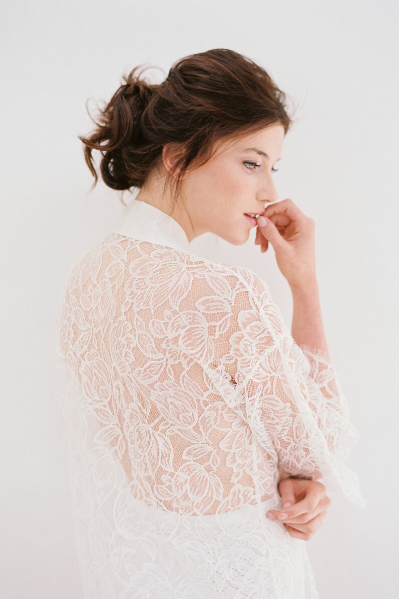 Op de afbeelding: Een witte kanten robe met een bloemenpatroon. De robe is doorschijnend en heeft lange mouwen. Het model draagt de robe en heeft haar haar in een knotje.