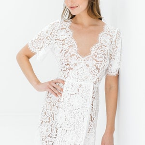 May include: A white lace mini dress with a V-neckline and short sleeves. The dress is made of delicate lace with a floral pattern.