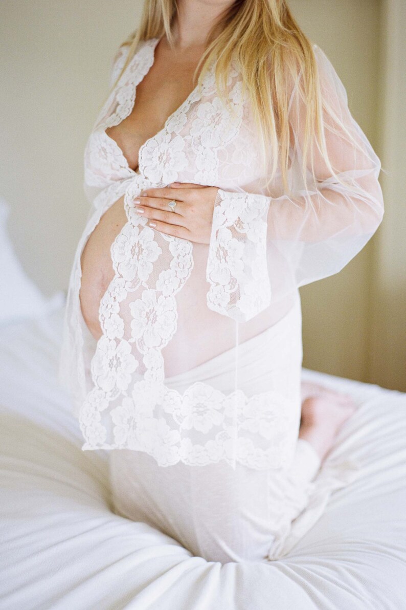 May include: A pregnant person wearing a white lace robe with a sheer overlay. The person is holding their belly with one hand and wearing a ring on their finger.