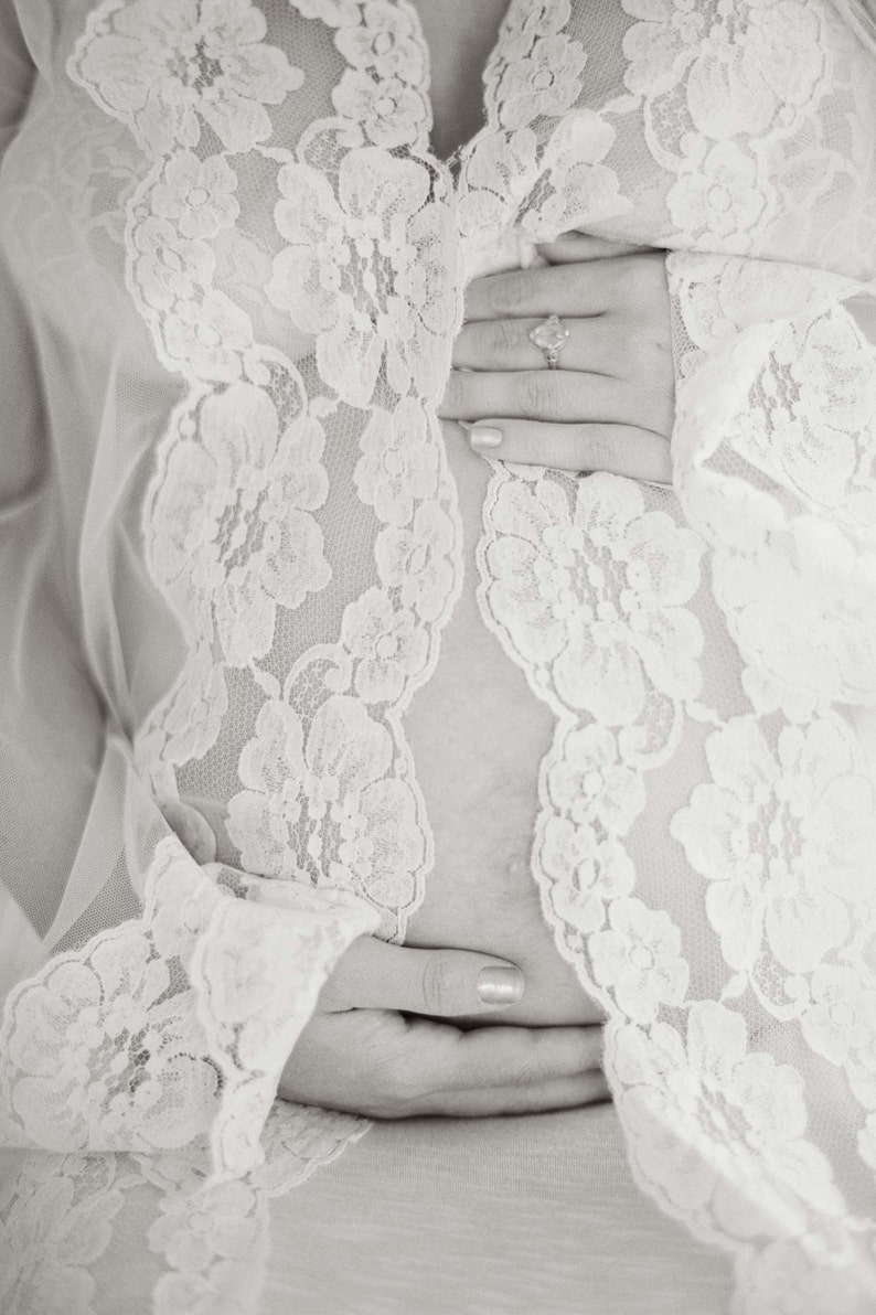 May include: A close-up of a pregnant woman's belly, covered in a white lace robe. The woman is holding her belly with both hands.