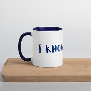 May include: White ceramic mug with a navy blue interior and handle. The mug features the phrase "I KNOW" in navy blue, handwritten-style lettering. The mug is sitting on a wooden cutting board.
