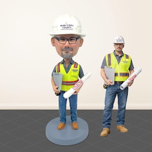 May include: A bobblehead figurine of a construction worker wearing a white hard hat with the text "MARICOPA COUNTY" and a yellow safety vest. The figurine holds a clipboard and a rolled blueprint marked "REJECTED". Another construction worker stands beside the bobblehead.