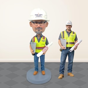 May include: A custom bobblehead figure of a person wearing a white Maricopa County Flood Control District hard hat, a grey polo shirt, a yellow high-visibility safety vest, and blue jeans, holding a clipboard and a rolled document marked REJECTED.