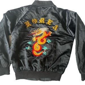 Puede incluir: Una chaqueta bomber de satén negro con un gran bordado detallado de un dragón amarillo y rojo en la espalda. Sobre el dragón, la palabra KOREA está bordada en letras amarillas. La chaqueta tiene cuello, puños y dobladillo acanalados.