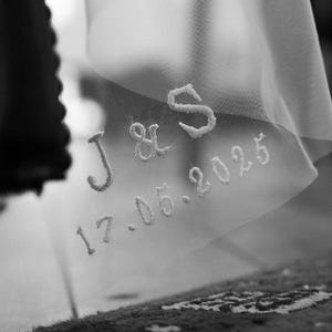 May include: Close-up of a sheer white fabric with embroidered text. The text includes the initials "J & S" and the date "17.05.2025". The fabric appears to be part of a wedding veil or similar bridal accessory.