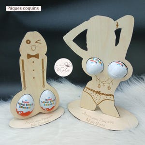 Peut inclure: Deux présentoirs en bois pour œufs de Pâques. L'un est en forme d'homme qui fait un clin d'œil avec un nœud papillon et des bretelles, l'autre est une silhouette de femme. Chacun contient deux œufs Kinder Surprise. Le texte sur la base dit "Joyeuses Pâques".