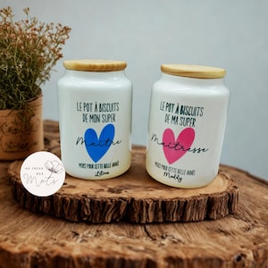 Peut inclure: Deux pots en céramique blancs avec des couvercles en bois, chacun avec du texte et un motif de cœur. L'un a un cœur bleu et le mot "Maitre", l'autre a un cœur rose et le mot "Maîtresse". Les pots sont sur une surface en bois.