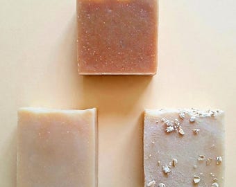 May include: Three rectangular bars of soap arranged in a triangular formation. The top bar is a medium brown colour with a textured top edge. The bottom left bar is a smooth, light beige colour. The bottom right bar is a pale cream with visible oats.