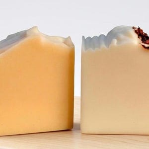 May include: Two handmade soap bars. One is a light orange color, the other is a pale cream color. The orange bar has a wavy top, and the cream bar has dried flower petals on top. Both are rectangular and sit on a wooden surface.