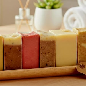 Könnte beinhalten: Ein Holztablett enthält sechs handgefertigte Seifenproben in verschiedenen Farben, darunter Gelb, Braun und Rot. Die Seifen haben strukturierte Oberflächen und ein Etikett mit der Aufschrift "Handcrafted Sample Soaps."