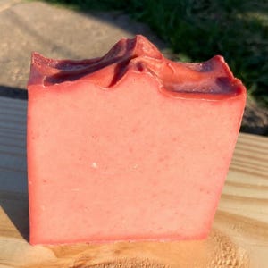 May include: A rectangular bar of handmade soap with a light pink hue. The top edge has a textured, uneven surface, while the sides are smooth. The soap is displayed on a wooden surface, showcasing its natural color and texture.