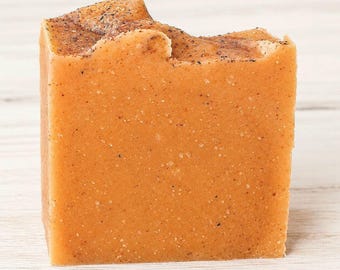 Op de afbeelding: Een rechthoekig stuk zeep met een warme oranje tint, voorzien van een getextureerd oppervlak met kleine donkerbruine deeltjes door het hele blok en een licht onregelmatige, rustieke bovenrand met een concentratie van donkere spikkels.