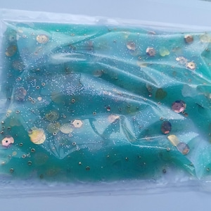 Op de afbeelding: Een doorzichtige plastic zak gevuld met een turquoise, gelachtige substantie. Goudkleurige en roze pailletten en glitter zijn door de gel verspreid. De zak is verzegeld en lijkt een sensorisch speelgoed of knutselmateriaal te zijn.