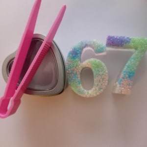 以下が含まれることがあります： ピンク色のプラスチック製のピンセットが、銀色のハート型の缶の隣に置かれています。「6」と「7」の数字は、青、緑、白の色合いの虹色のビーズで飾られています。オブジェクトは白い表面にあります。