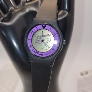 Puede incluir: Un reloj negro de Disney Channel con bisel y esfera morados. La esfera del reloj presenta la palabra "CAPTAIN" y una silueta de Mickey Mouse. La correa del reloj es negra.