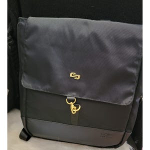 May include: Black backpack with a flap closure and a gold-colored clasp. The backpack has a gold-colored logo on the front and a gray bottom panel. The brand name "solo NEW YORK" is printed on the bottom.