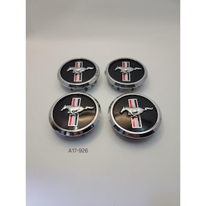 May include: Four black and silver hubcaps with the Mustang logo. Each hubcap features a silver running horse emblem on a black background, with red and white vertical stripes. The text "A17-926" is visible.