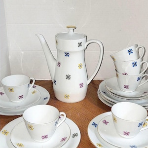Könnte beinhalten: Ein weißes Keramik-Teeservice mit Blumenmuster. Das Set beinhaltet eine hohe Kaffeekanne, Teetassen und Untertassen. Die Blumen sind in Blau-, Gelb-, Grau- und Lilatönen gehalten. Die Ränder der Tassen und Untertassen haben einen goldenen Rand.