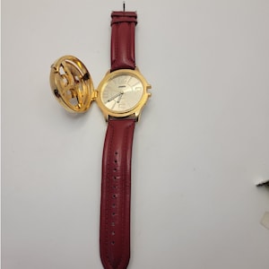 May include: A gold-toned wristwatch with a burgundy leather strap. The watch face is gold with black numerals and hands. The watch has a hinged cover with a decorative design. The strap is approximately 23 cm long.