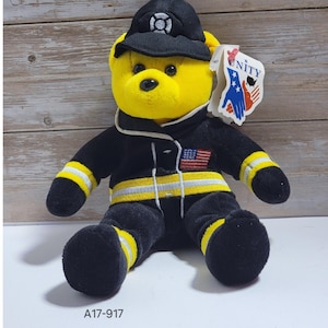 May include: A yellow teddy bear wearing a black firefighter uniform with yellow and silver stripes. The bear has a black hat and an American flag patch. A tag that says "UNITY" is attached to the bear.