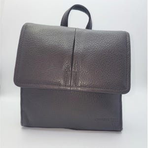 Puede incluir: Una mochila de cuero gris oscuro con cierre de solapa y asa superior. La mochila tiene una superficie texturizada y la marca "KENNETH COLE" es visible en la esquina inferior derecha.