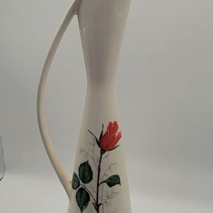 May include: Cream-colored ceramic pitcher with a tall, slender design and a curved handle. A red rose with green leaves is painted on the front. The pitcher is approximately 12 inches tall.