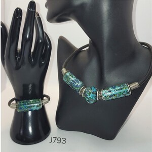 Könnte beinhalten: Ein Halsketten- und Armband-Set mit zylindrischen und kugelförmigen Perlen mit blau-grünen und weißen Mustern. Die Perlen sind mit silberfarbenem Metall akzentuiert und auf einer schwarzen Kordel und einem Armbandband aufgefädelt. Der Schmuck wird auf schwarzen Schaufensterpuppen präsentiert.