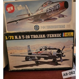 Op de afbeelding: Twee modelvliegtuigdozen. De bovenste doos toont een zilveren en rode F-84 Thunderstreak jet met de tekst "FS-713". De onderste doos toont een zilveren en zwart N.A.T-28 Trojan/Fennec propeller vliegtuig. Beide dozen zijn voorzien van schaalinformatie.