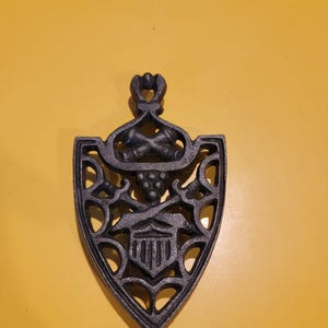 May include: A black cast iron trivet with a shield shape. The trivet features an ornate design with a central emblem and decorative cutouts. The handle has a heart-shaped design. The trivet is designed to protect surfaces from hot cookware.