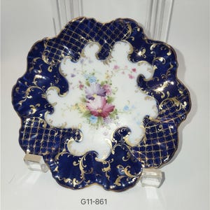 May include: A decorative porcelain plate with a scalloped edge. The plate features a dark blue border with a gold lattice pattern and gold floral accents. The center has a white background with a floral bouquet.