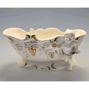 Könnte beinhalten: Eine weiße dekorative Keramikschale mit goldenen Akzenten. Die Schale zeigt auf einer Seite einen Engel und goldene Weinreben-Designs. Die Schale hat einen gewellten Rand und steht auf einem kleinen Sockel. Ein dekoratives Stück für das Zuhause.