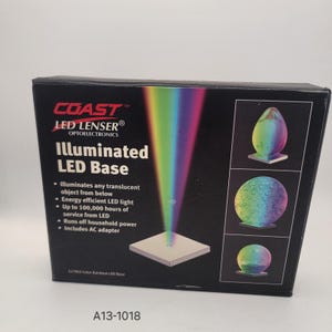 Op de afbeelding: Een zwarte doos voor een verlichte LED-basis. De doos toont het COAST LED LENSER logo en productdetails. De doos toont een regenbooglichteffect en voorbeelden van verlichte objecten. De tekst "Illuminated LED Base" is prominent aanwezig.