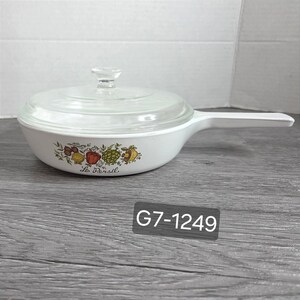 Pode incluir: Uma frigideira de cerâmica branca com cabo longo e tampa de vidro transparente. O exterior apresenta uma ilustração botânica colorida de vegetais e o texto Le Persil. A frigideira tem o código alfanumérico G7-1249 na lateral.
