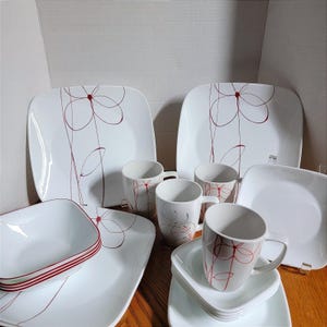 May include: White dinnerware set with a red floral design. The set includes square plates, bowls, and mugs. The bowls have a red stripe around the rim. The mugs and plates feature a stylized flower pattern.