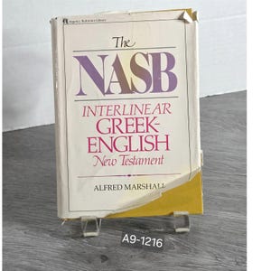 May include: A hardcover book titled The NASB Interlinear Greek-English New Testament by Alfred Marshall. The cover is off-white with purple and red text, featuring a torn dust jacket at the bottom right corner and a yellow spine edge.