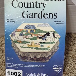 Puede incluir: Un kit de punto de cruz titulado "Country Gardens" con una caja hexagonal con una escena campestre bordada. La caja mide aproximadamente 15 cm de diámetro y tiene un ribete de encaje. El kit incluye materiales de construcción e instrucciones.