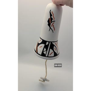 Puede incluir: Campana de cerámica blanca con diseños geométricos negros y color óxido. La campana tiene una forma cilíndrica larga con una base acampanada. Una cuerda con un pequeño tirador gris en forma de estrella cuelga de la parte inferior. El texto "G8-533" es visible.