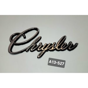 Puede incluir: Un emblema Chrysler cromado con una fuente cursiva. El emblema tiene un contorno negro y está sobre un fondo blanco. Una pequeña pegatina gris con el texto "A13-527" está debajo.
