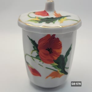 May include: White ceramic jar with a lid, adorned with a vibrant red poppy floral design. The lid and rim feature gold trim. The jar has a cylindrical shape and a small handle on the lid. The text "G8-578" is visible on the bottom right.