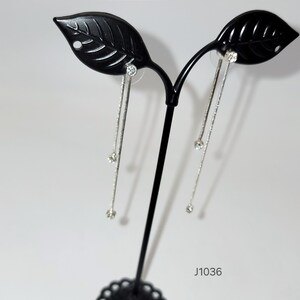 May include: Silver dangle earrings on a black leaf-shaped display stand. Each earring has a fine chain with a small, sparkling crystal. The earrings are presented against a white backdrop.