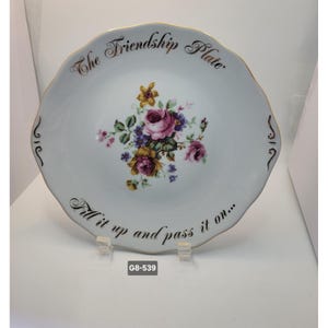 May include: White ceramic plate with a scalloped edge and gold trim. The plate features a floral bouquet of pink, purple, and yellow flowers. Text reads "The Friendship Plate" and "Fill it up and pass it on..."