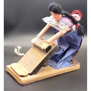 May include: A handcrafted doll depicting a woman in traditional attire, kneeling and using a wooden washboard. The doll features a purple blouse with a white lace collar, a blue skirt, and a long black braid with red and white accents. The washboard is made of wood.