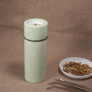 May include: A light green cylindrical container with a black band, next to a small white oval dish filled with brown dried herbs. A pair of silver tweezers rests nearby. The container has a white interior.