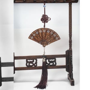 Puede incluir: Un abanico plegable de madera con intrincados diseños recortados está suspendido de un expositor de madera marrón oscuro. Una borla decorativa cuelga debajo del abanico. El soporte tiene diseños recortados decorativos.